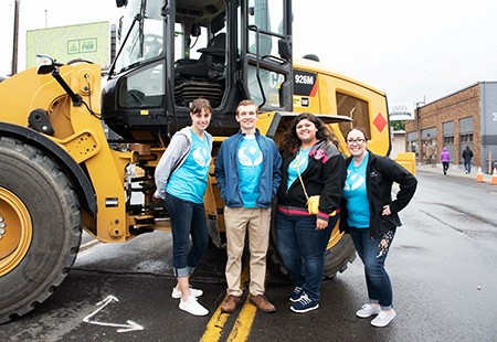 Four Numerica employees, one man and three women, wearing blue t-shirts, jackets and jeans, standing in front of a wheel loader on Monroe Street in Spokane. People walking down sidewalk further down the street. 