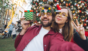 Hipster couple in front of a Christmas tree holding a present and wearing fun Christmas glasses