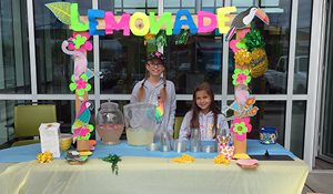 Two young girls stand in front of a Numerica Credit Union branch selling lemonade at a Luau decorated lemonade stand