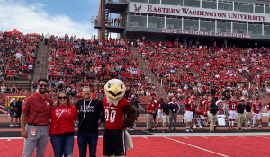 Group of three people standing next to the Eastern Washington University mascot on a red football turf in front of bleachers filled with attendees sitting.