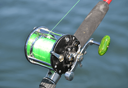 Fishing reel with grey grip, green line, silver and green crank and red pole. Lake or river water in the background. 