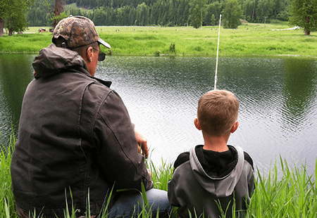 Man in brown jacket, camouflage hat and sunglasses with young boy, blond hair and grey and black jacket. Boy holding a fishing rod with fishing line dropped into lake. Grassy field and pine trees in the background. Tall grasses in the foreground. 