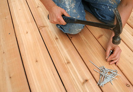 Person kneeling on wooden deck in paint speckled jeans hammering nails into a new deck with a pile of nails next to their hand. 