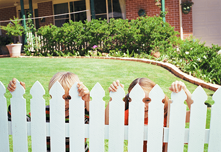 Two children hiding behind white picket fence in a yard. Green grass with shrubs bordering a brick house. 