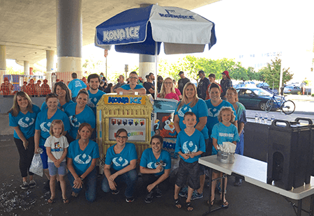 Group of people, children and adults, in blue Numerica logo t-shirts standing next to a yellow ice cream stand under a blue umbrella that says Kona Ice. 