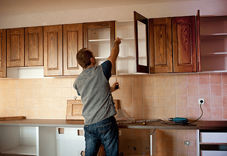 Man in grey vest and blue jeans measuring cabinets in a kitchen that is being remodeled. Dark wooden cabinets, tile backsplash. 