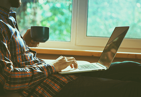 Man in plaid shirt drinking coffee and working on a laptop. Window, trees in the foreground. 