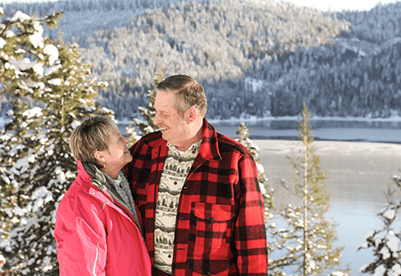 Man in red and black plaid jacket and cream colored shirt hugging woman in pink coat and grey fleece. Man and woman are standing outside with a lake and pine trees and a mountain in the background. 