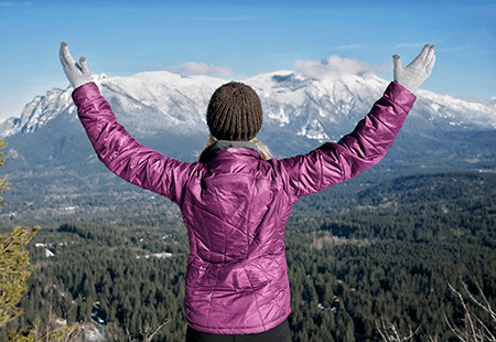 Woman in a purple coat and brown knit cap, white gloves, holding her hands up and looking out over pine trees and white capped mountain in the distance. 