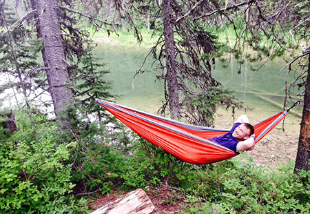 Man in blue shirt laying in a red hammock tied between two pine trees. Shallow river in the background. 