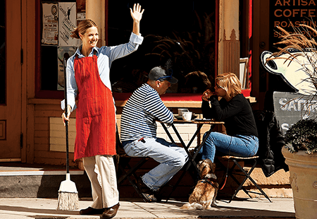 Woman in a denim shirt, tan pants and a red apron carrying a broom and waving outside on sidewalk in front of a coffee shop. Two patrons sitting at table drinking coffee. 