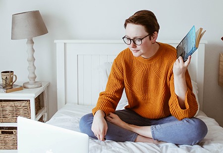 Girl with short hair wearing black glasses, orange sweater and blue jeans, sitting on a bed looking at laptop, holding book.