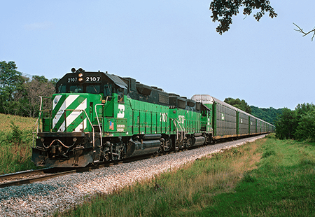Freight train pulled by two green engine cars pulling several silver freight cars on a track running through green field with trees surrounding train track and field.