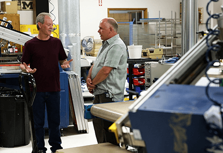 Two older men talking in a manufacturing shop. One man is in all black and the other is in a plaid shirt. Manufacturing equipment and machines is scattered around them. 