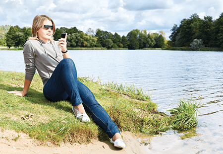 Woman with blond hair and sunglasses in grey shirt and jeans sitting on at a lakefront while looking at her cell phone. Trees and lake in the background. 