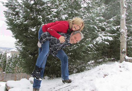 Mature man in grey and black flannel and jeans, grey hair, carrying and playing in the snow with blond woman in red shirt and blue jeans. 