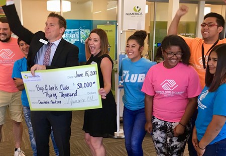 Man in black suit holding a large Numerica Credit Union check for Boys and Girls Club Richland, WA. Group of kids and Numerica employees celebrating, smiling, cheering. 