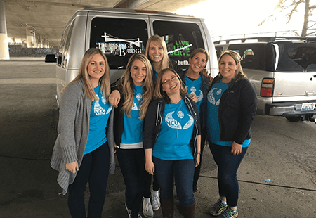 6 women in Numerica blue volunteer shirts and jeans standing at the rear of a silver van with "Blessings under the Bridge" logo and Live, love, serve window decals.