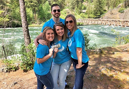 Four Numerica employees, one man and three women, in light blue t-shirts and jeans, standing at the edge of a river  with rapid. Bridge over the river and pine trees in the background.